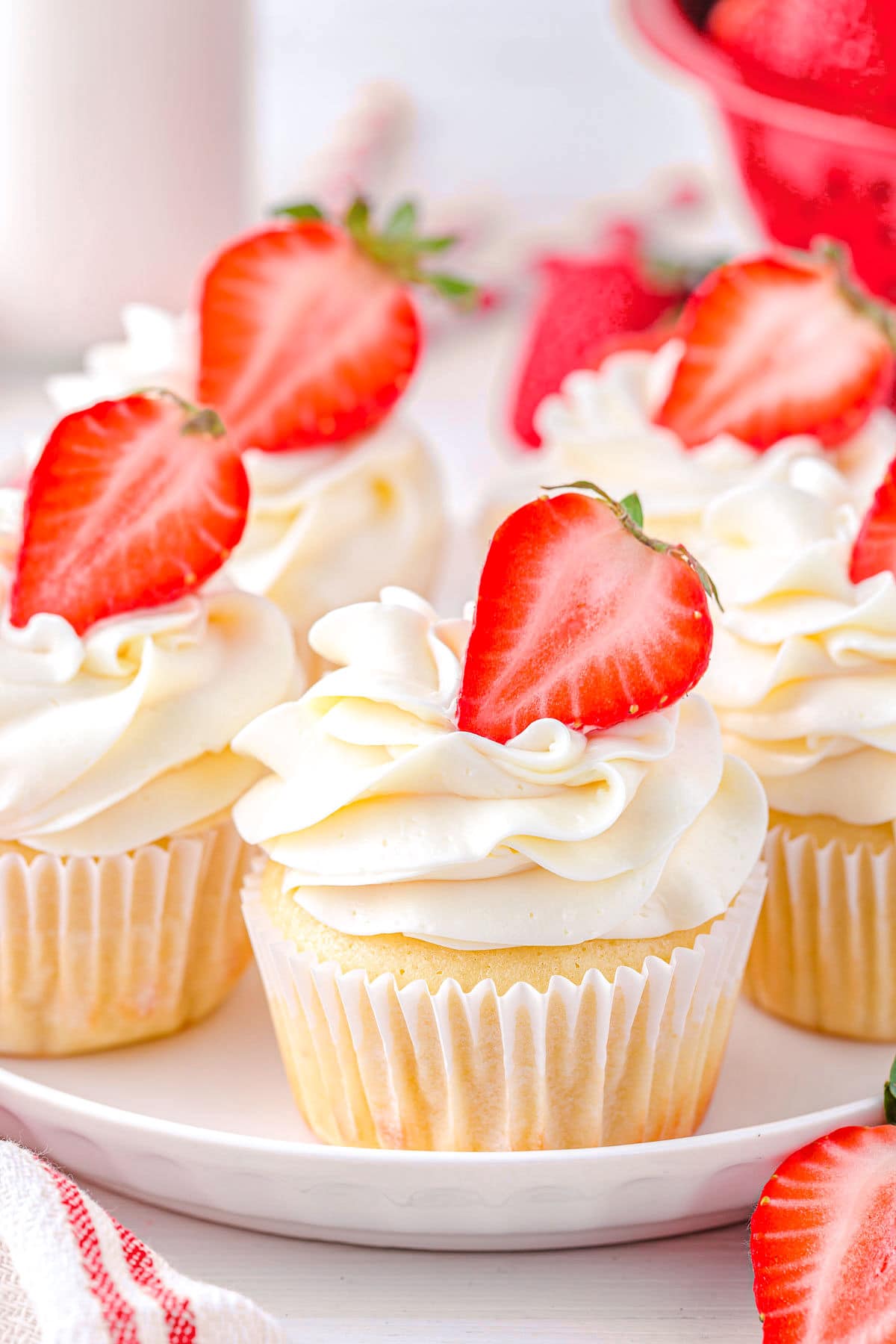 five strawberry shortcake cupcakes on a white plate topped with strawberry halves and buttercream frosting.