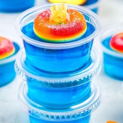 three blue jello shots stacked with lids. top shot has a peach ring and gummy bear on it.