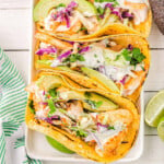 top down view of four fish tacos on a white rectangle plate with avocados and cilantro and lime wedges around the plate.