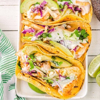 top down view of four fish tacos on a white rectangle plate with avocados and cilantro and lime wedges around the plate.