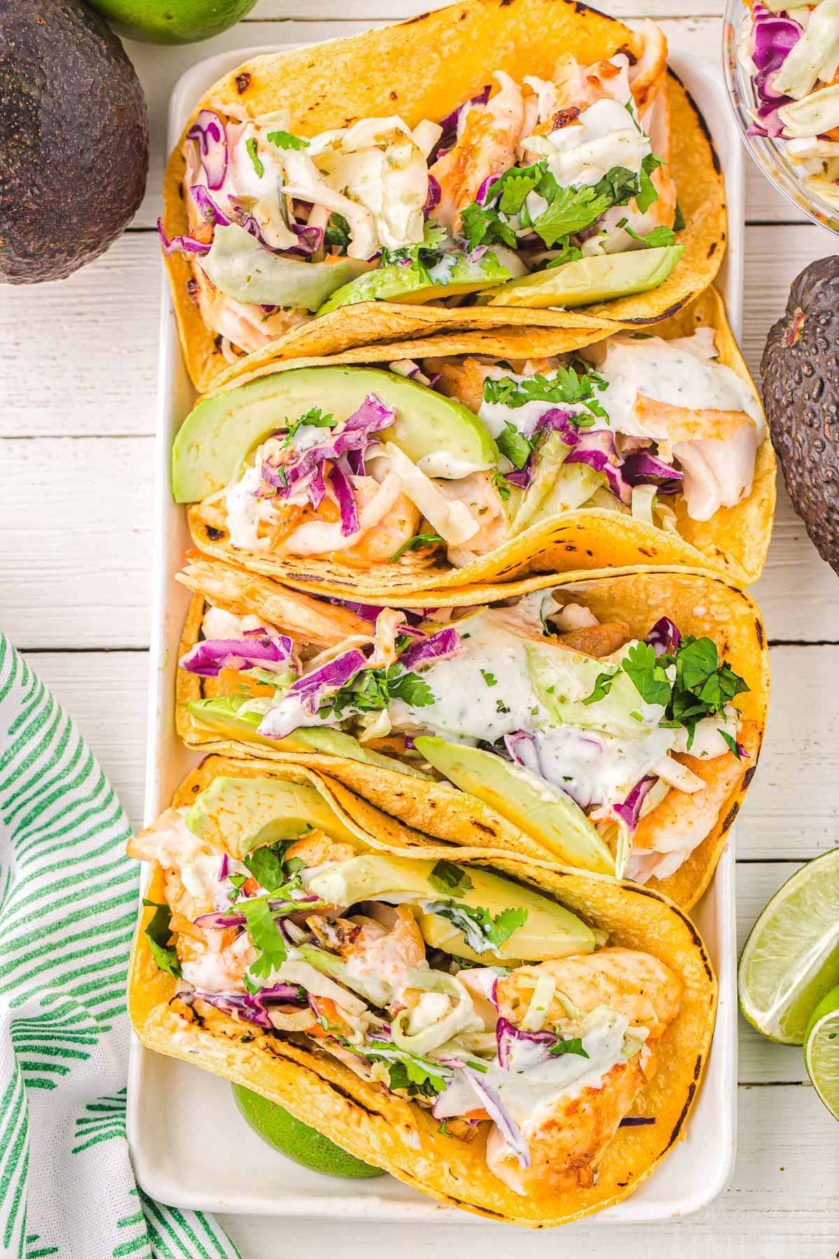 top down view of four fish tacos on a white rectangle plate with avocados and cilantro and lime wedges around the plate.