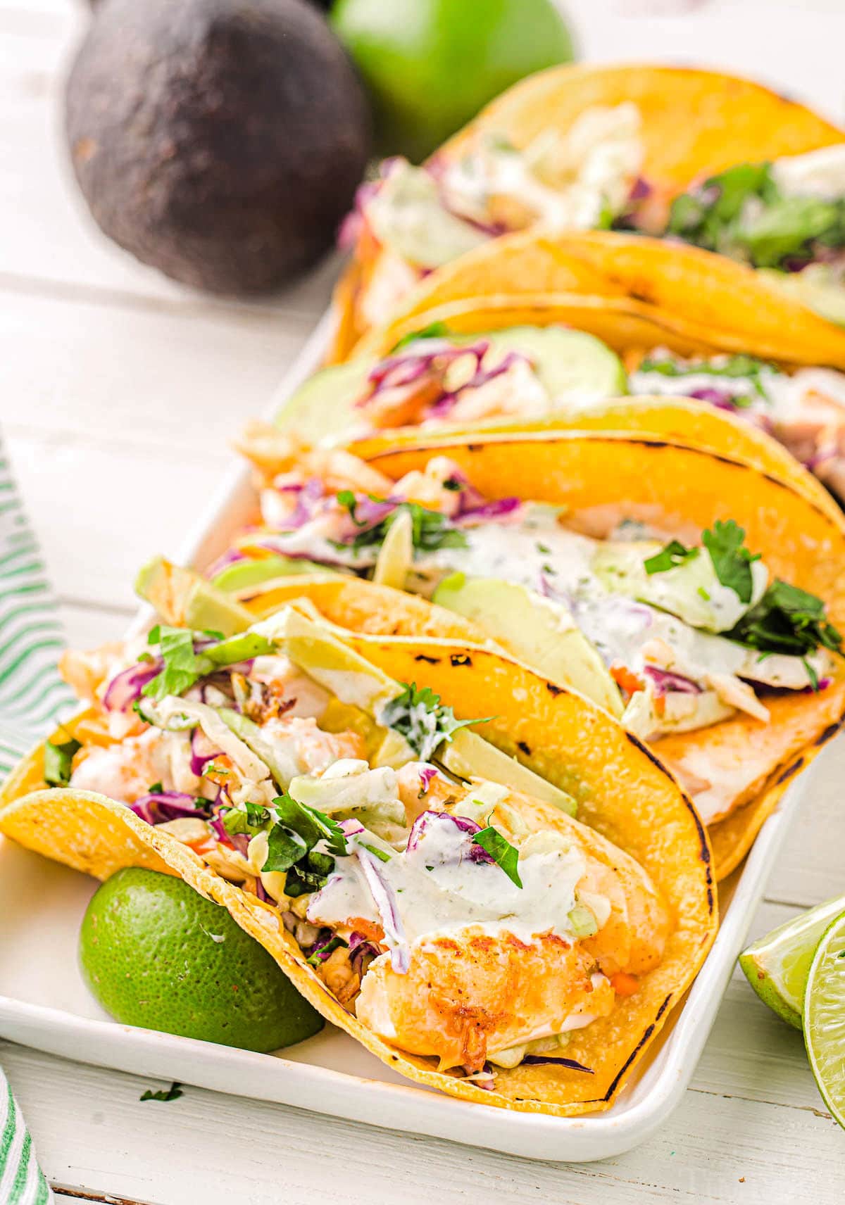 three fish tacos on a white platter topped with fish taco sauce and cilantro. lime wedges are placed next to the tacos. an avocado can be seen in the background.