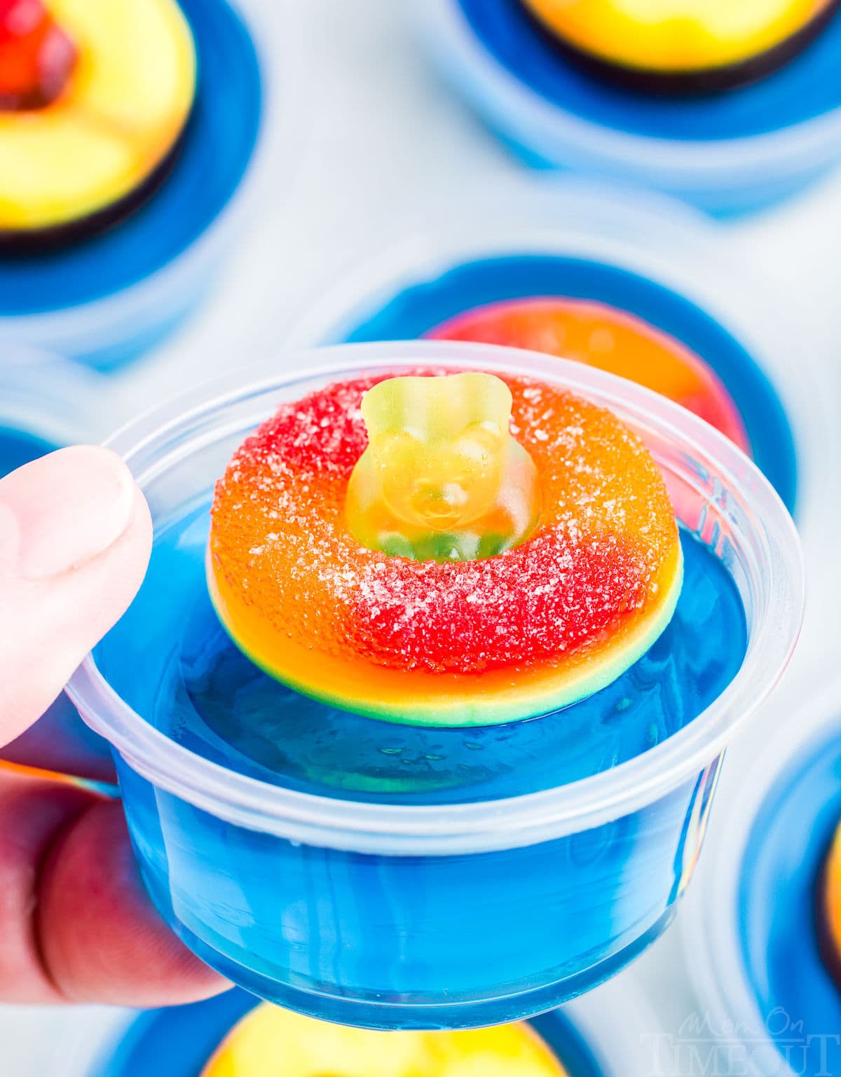 hand holding single blue jello shot with a peach ring and gummy bear on top. more jello shots can be seen in the background.