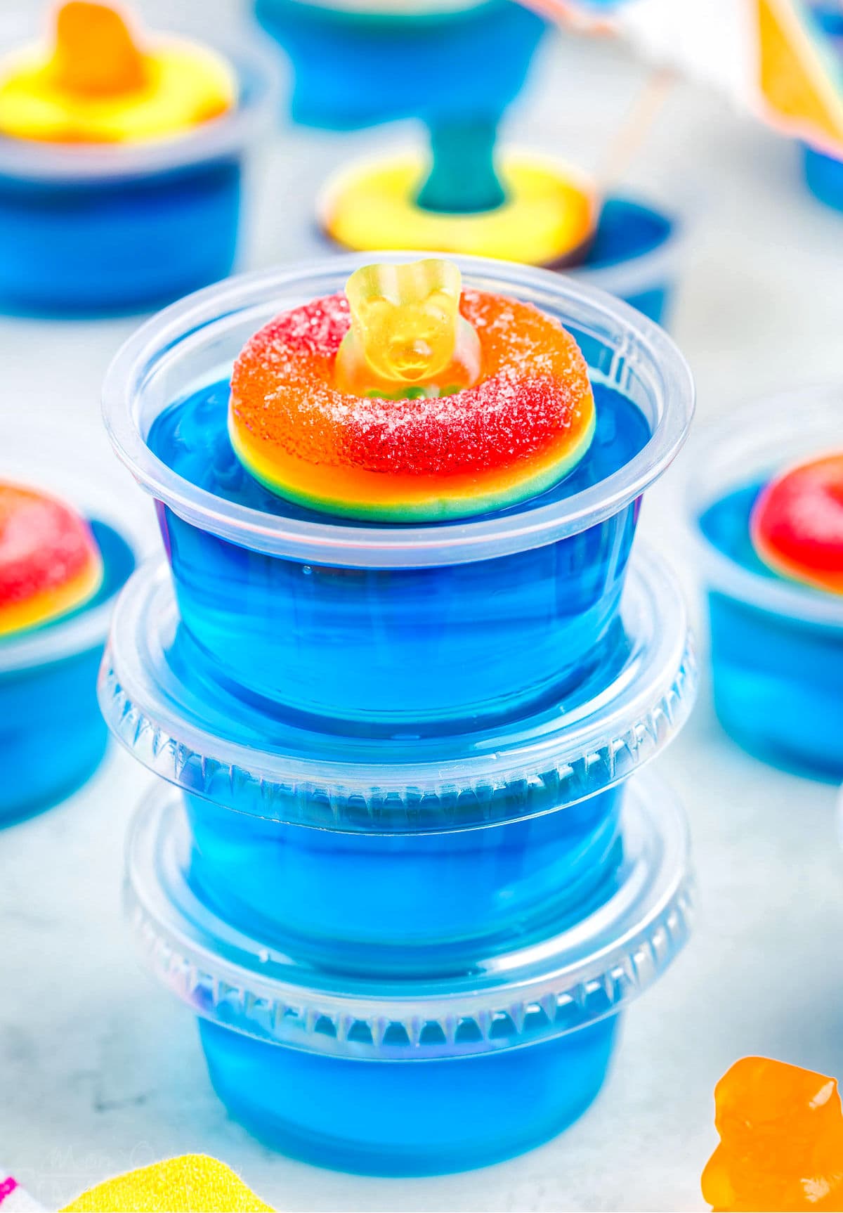 three blue jello shots stacked with lids. top shot has a peach ring and gummy bear on it.