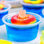 blue pool party jello shot with gummi bear sitting on white surface. more jello shots can be seen in the background.