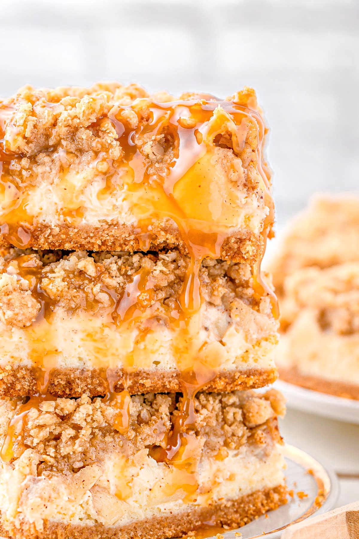 three caramel apple cheesecake bars stacked on a white plate with a gold rim. caramel sauce is drizzled on top of the bars.