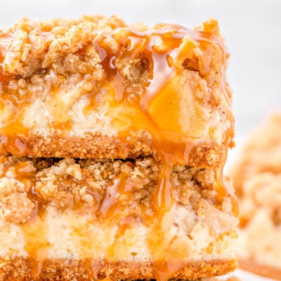 two caramel apple cheesecake bars stacked on a white plate with a gold rim. caramel sauce is drizzled on top of the bars.