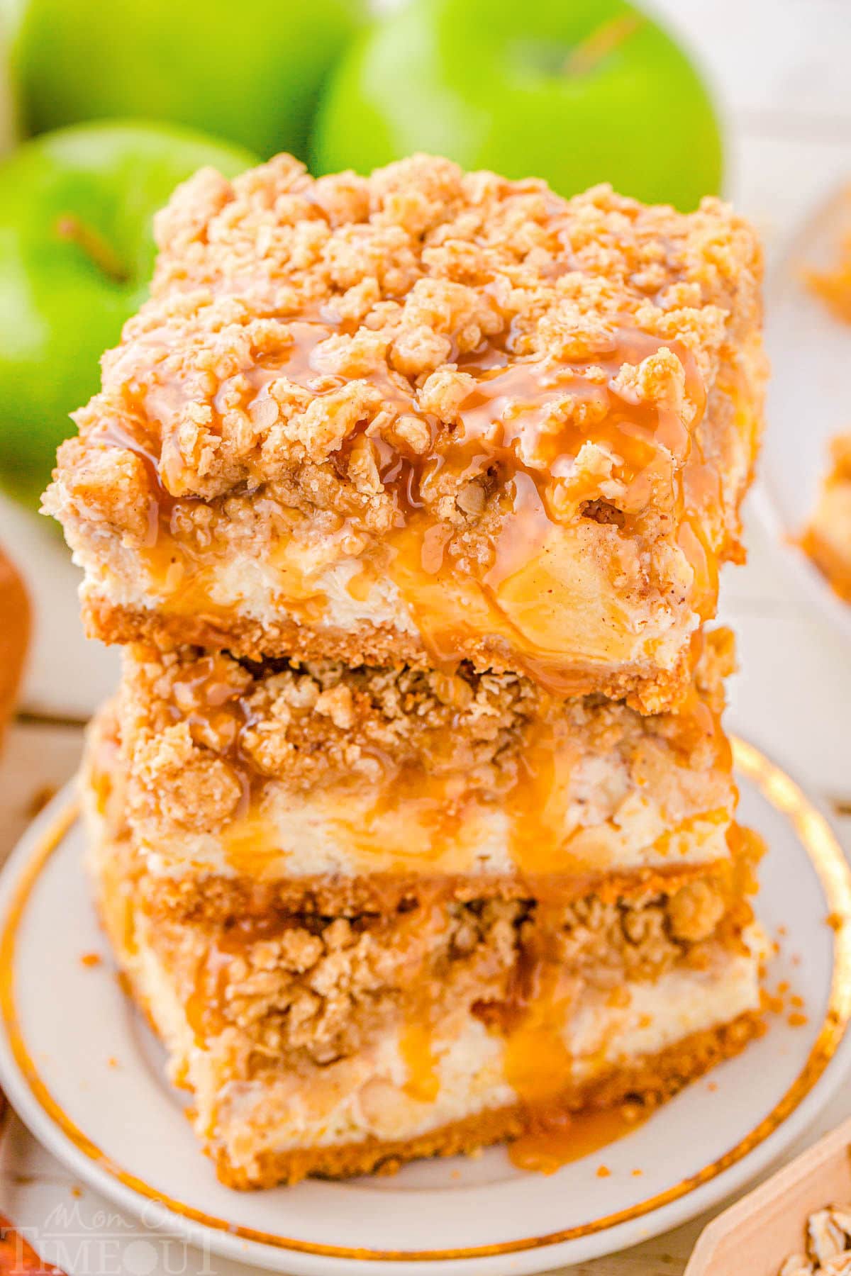 three caramel apple cheesecake bars stacked on a white plate. the entire stack has caramel drizzled on top of it.