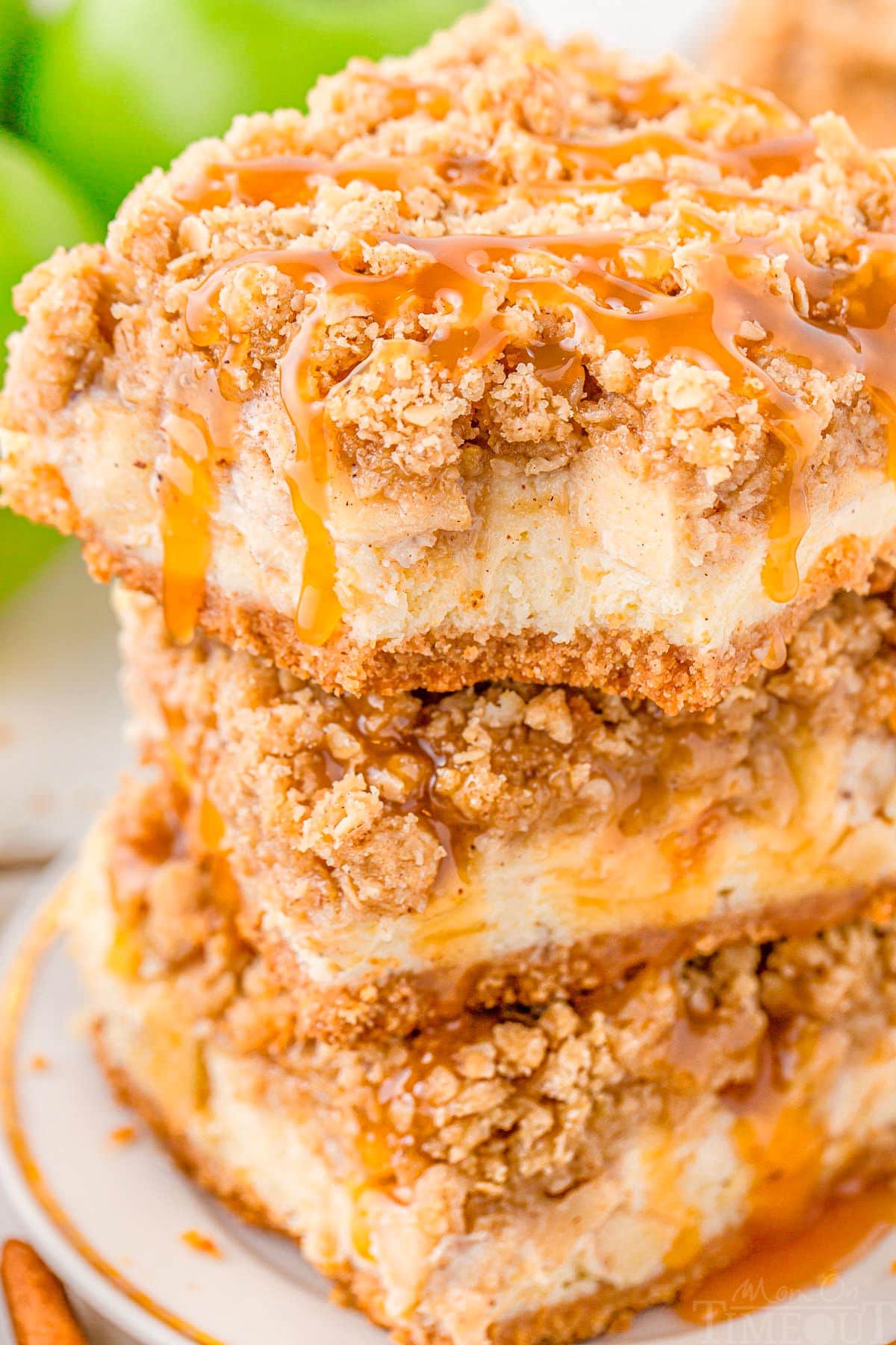 three caramel apple cheesecake bars stacked on a white plate. the top bar has a bite taken out of it and the entire stack has caramel drizzled on top of it.