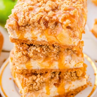 three caramel apple cheesecake bars stacked on a white plate. the entire stack has caramel drizzled on top of it.