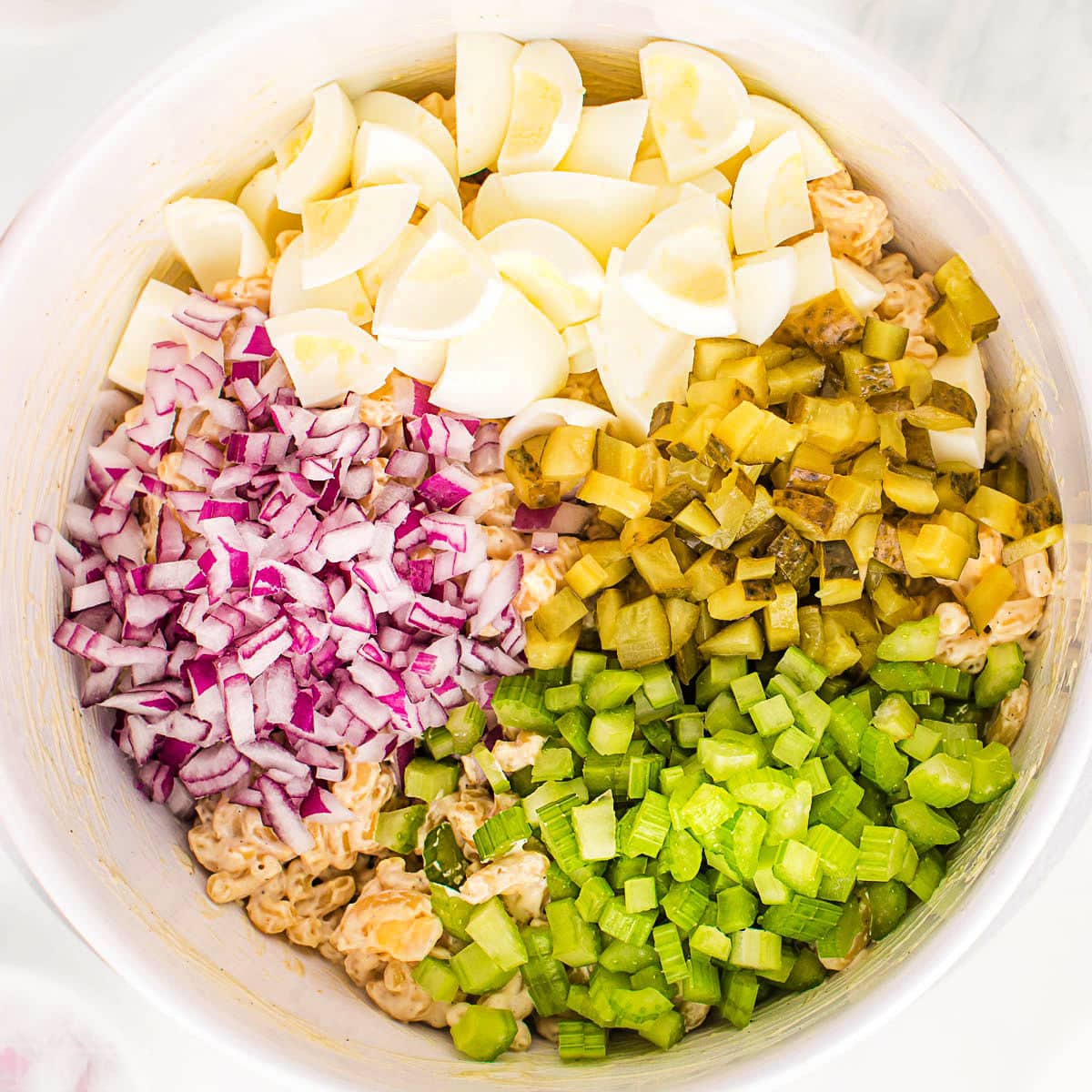 top down view of a white bowl filled with macaroni salad with eggs, pickles, celery, and red onion on top waiting to be stirred in.
