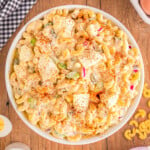 top down view of deviled egg macaroni salad in large white serve bowl sitting on wood backdrop. eggs and hard boiled eggs are around the bowl along with a white and black checked towel.