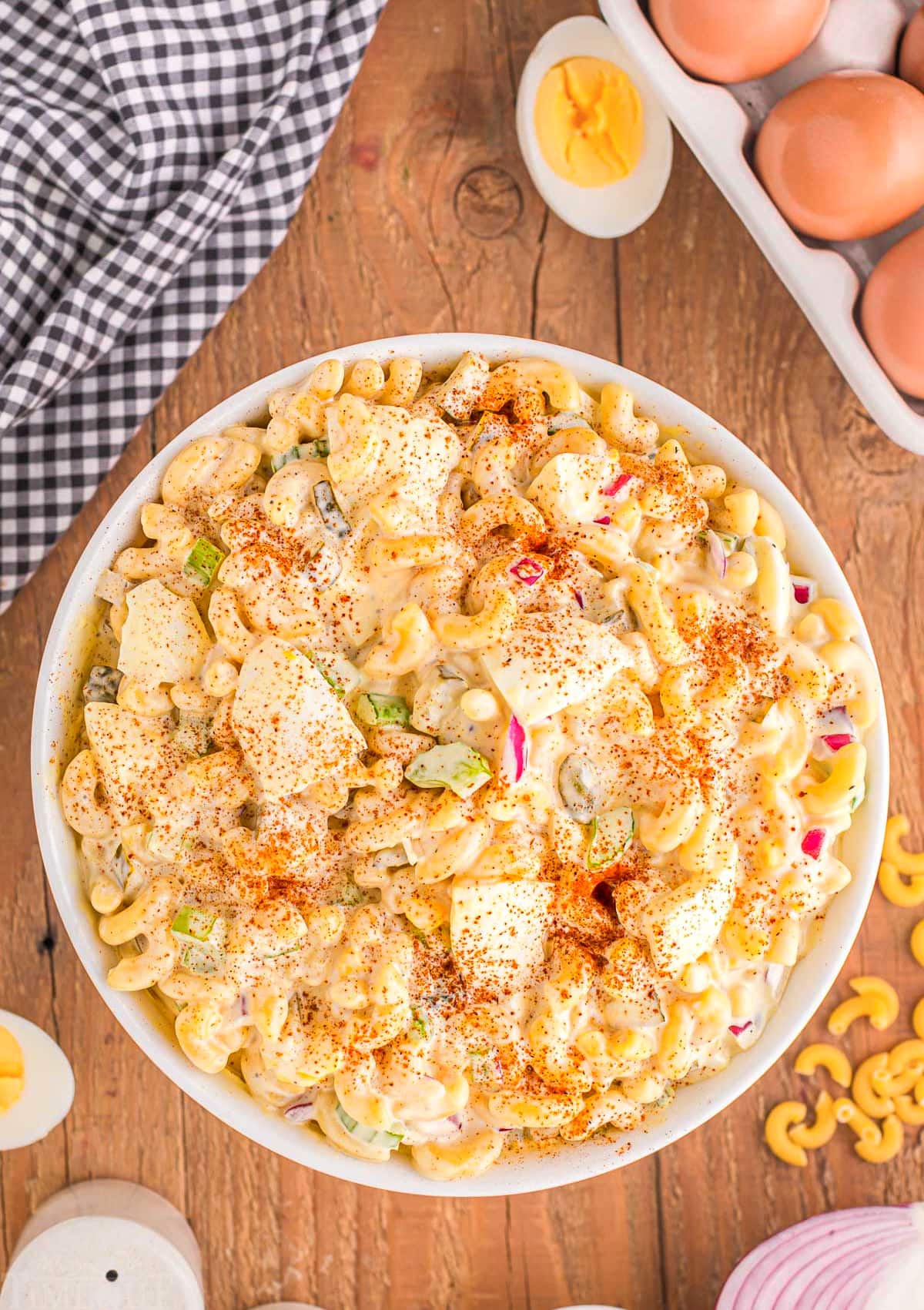 top down view of deviled egg macaroni salad in large white serve bowl sitting on wood backdrop. eggs and hard boiled eggs are around the bowl along with a white and black checked towel.