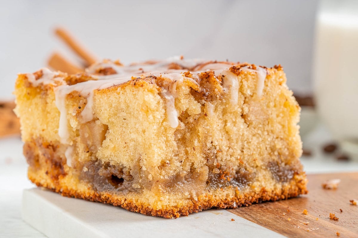 one piece of apple coffee cake sitting on a marble and wood cutting board. coffee cake has a little glaze drizzled over the top.