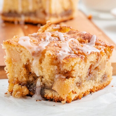 one piece of apple coffee cake sitting in front of a cutting board with three more pieces stacked together. coffee cake has a little glaze drizzled over the top.