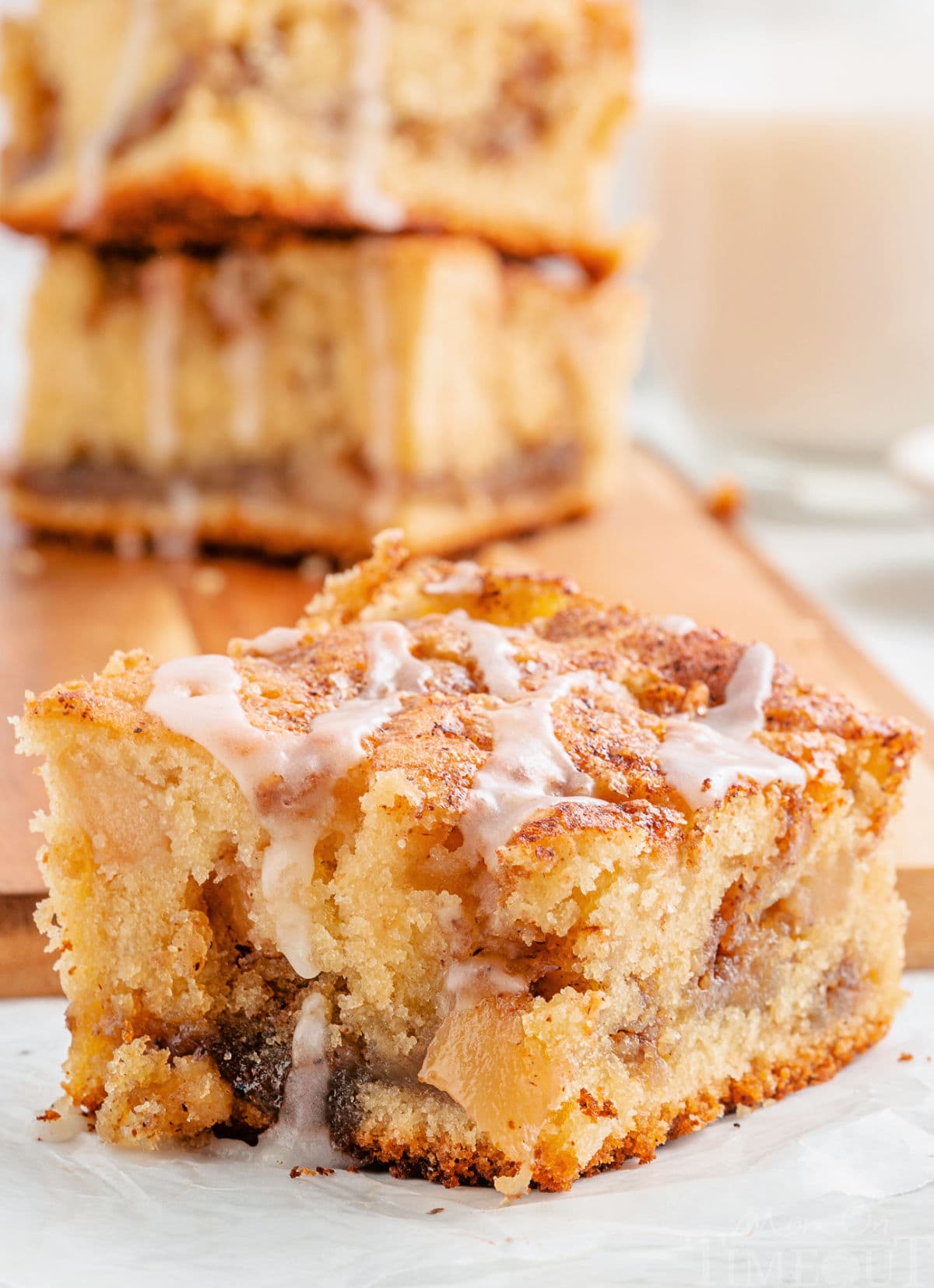 one piece of apple coffee cake sitting in front of a cutting board with three more pieces stacked together. coffee cake has a little glaze drizzled over the top.