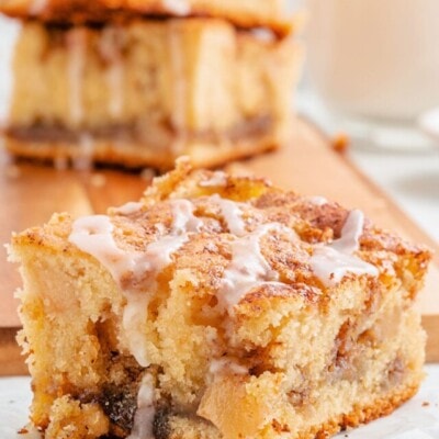 one piece of apple coffee cake sitting in front of a cutting board with three more pieces stacked together. coffee cake has a little glaze drizzled over the top.