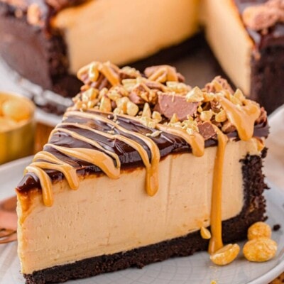 single slice of peanut butter cheesecake sitting on small round white plate. cheesecake is topped with peanut butter topping and reese's peanut butter cups chopped up.