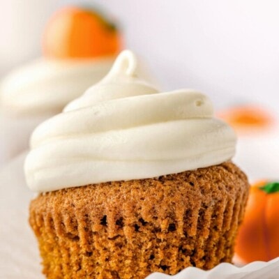 single pumpkin cupcake with cream cheese frosting piped on top and cupcake liner mostly pulled away from the cupcake itself. more cupcakes can be seen in the background.