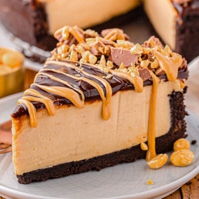 single slice of peanut butter cheesecake sitting on small round white plate. cheesecake is topped with peanut butter topping and reese's peanut butter cups chopped up.