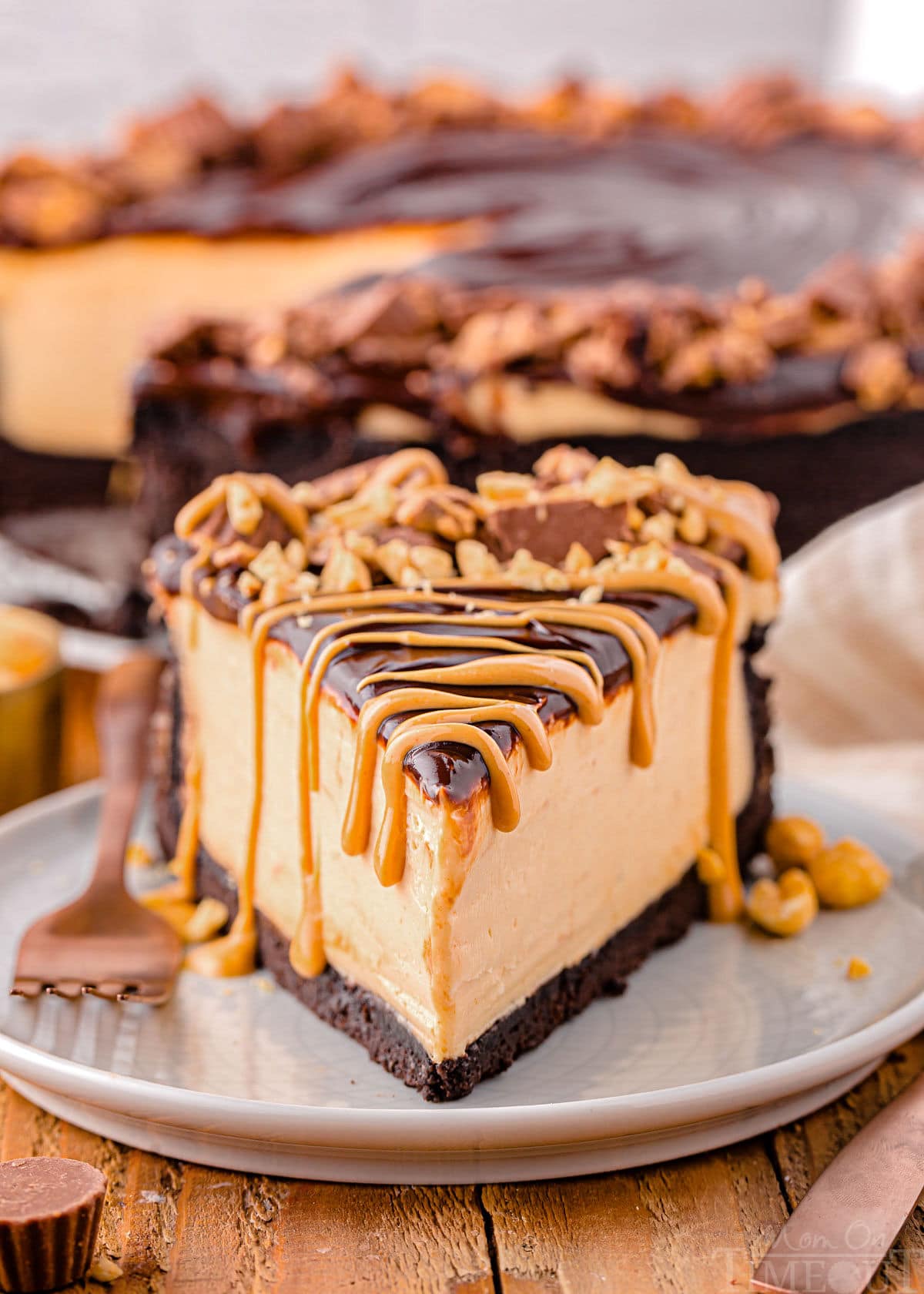 single slice of peanut butter cheesecake sitting on small round white plate. cheesecake is topped with peanut butter topping and reese's peanut butter cups chopped up.