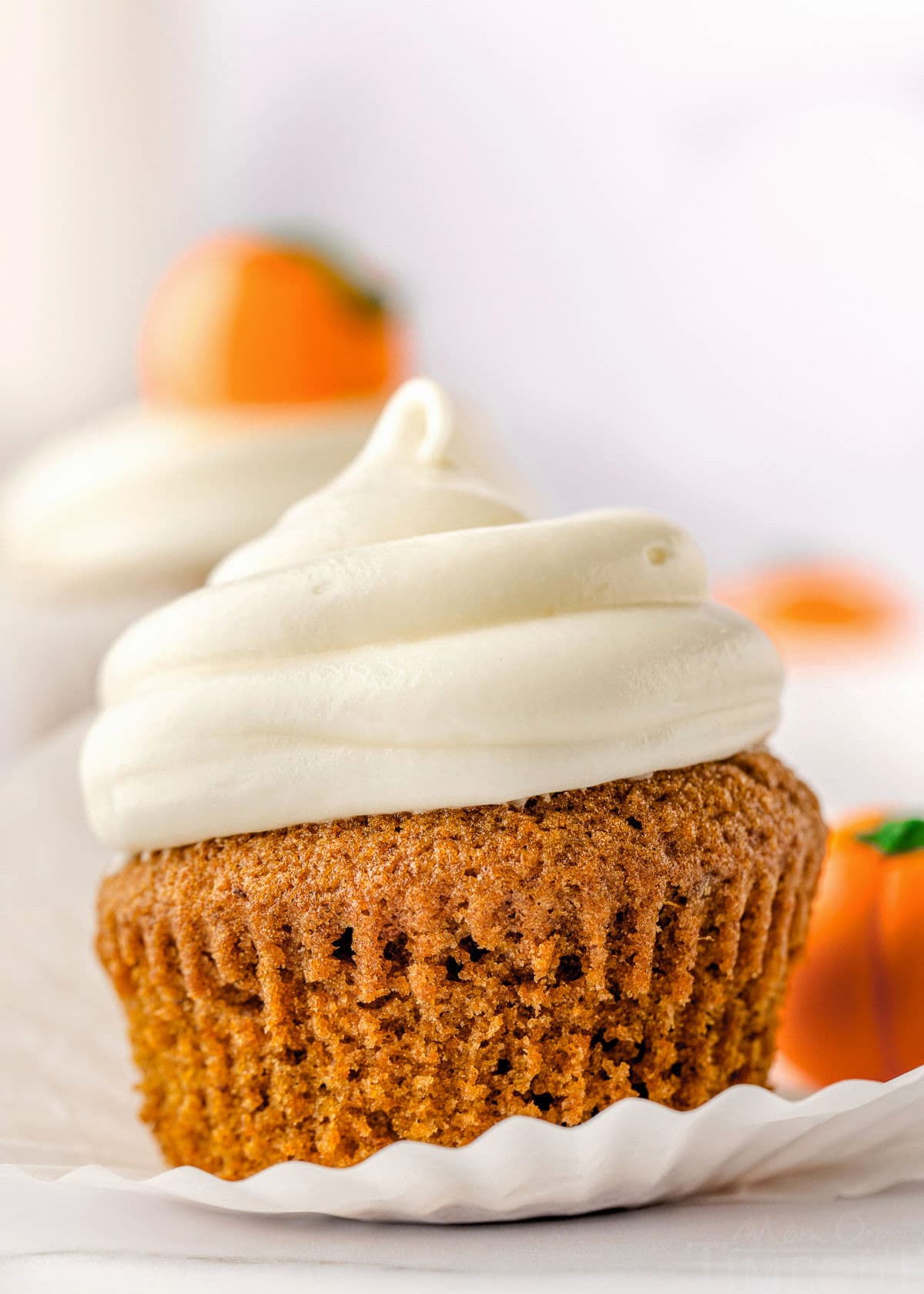 single pumpkin cupcake with cream cheese frosting piped on top and cupcake liner mostly pulled away from the cupcake itself. more cupcakes can be seen in the background.