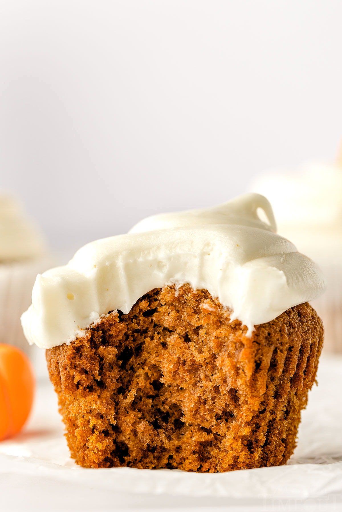 single pumpkin cupcake sitting on white cupcake liner frosted with cream cheese frosting. one large bite has been taken from the cupcake revealing a tender crumb.