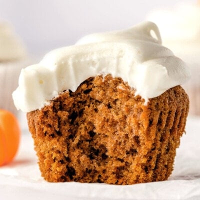 single pumpkin cupcake sitting on white cupcake liner frosted with cream cheese frosting. one large bite has been taken from the cupcake revealing a tender crumb.