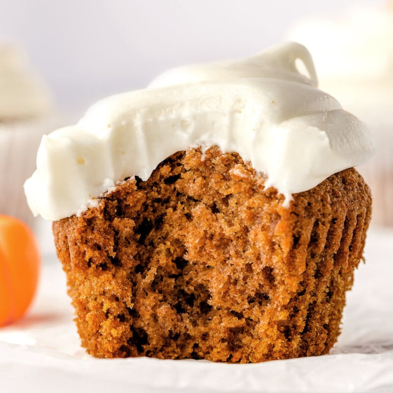single pumpkin cupcake sitting on white cupcake liner frosted with cream cheese frosting. one large bite has been taken from the cupcake revealing a tender crumb.