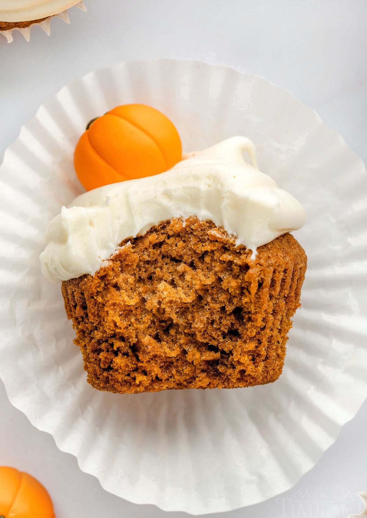 top down shot of pumpkin cupcake lying on it's side with a bite taken out showing the moist and tender crumb of the cupcake. the cupcake is sitting on it's white liner and is topped with a fondant pumpkin.