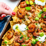 shrimp nachos on a sheet tray with lots of topping. hand pulling a loaded chip off of the nachos.
