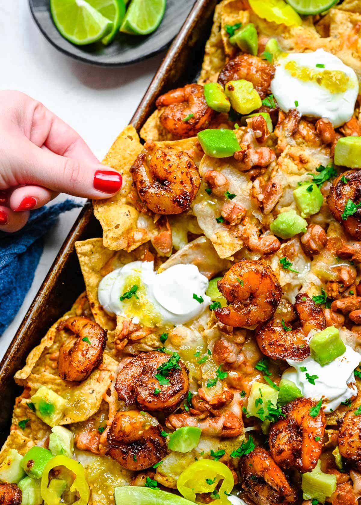 shrimp nachos on a sheet tray with lots of topping. hand pulling a loaded chip off of the nachos.