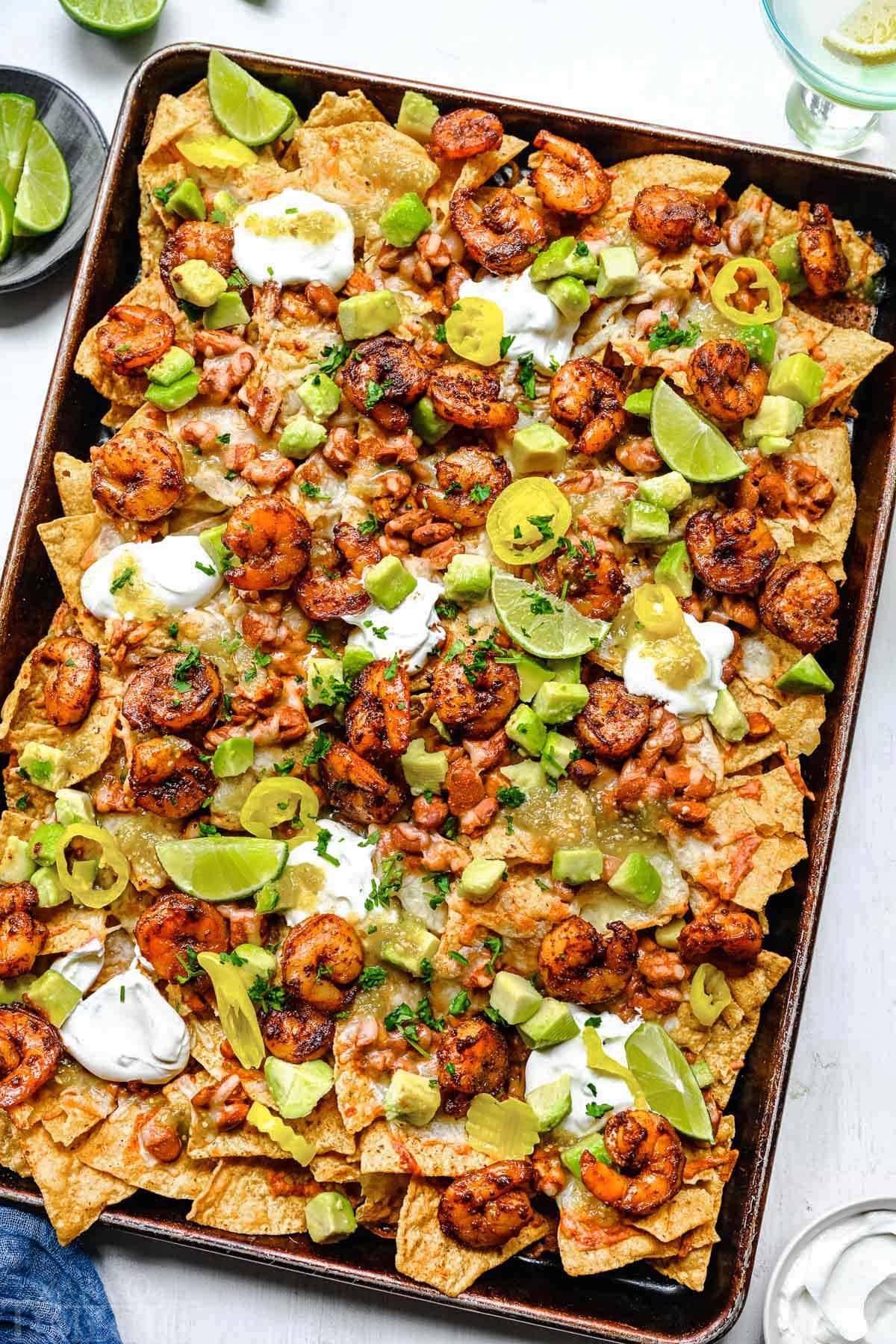 top down look at shrimp nachos prepared on a sheet pan topped with sour cream, avocados and salsa.