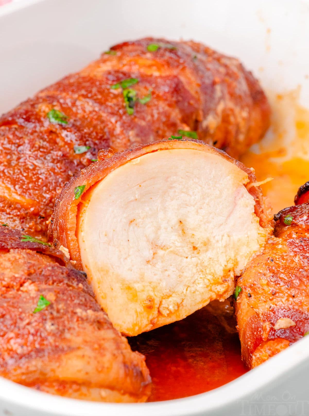 baking dish with bacon wrapped chicken. one chicken breast is cut in half. the chicken is topped with parsley.