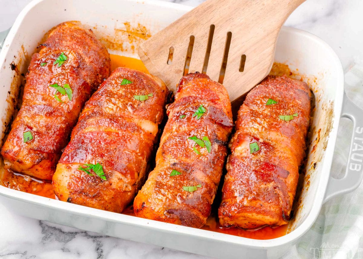 four bacon wrapped chicken breasts in staub baking dish with a wood server tucked under the end of one chicken breast.