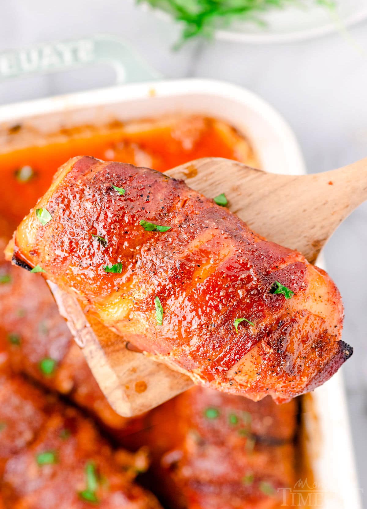 top down look at a casserole dish with bacon wrapped chicken breasts. one piece of chicken is being lifted out of the dish with a wood spoon.