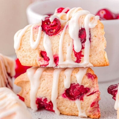 two small cranberry scones stacked with a simple glaze drizzled on top. a small white bowl of cranberries are in back as well as more scones to the sides.