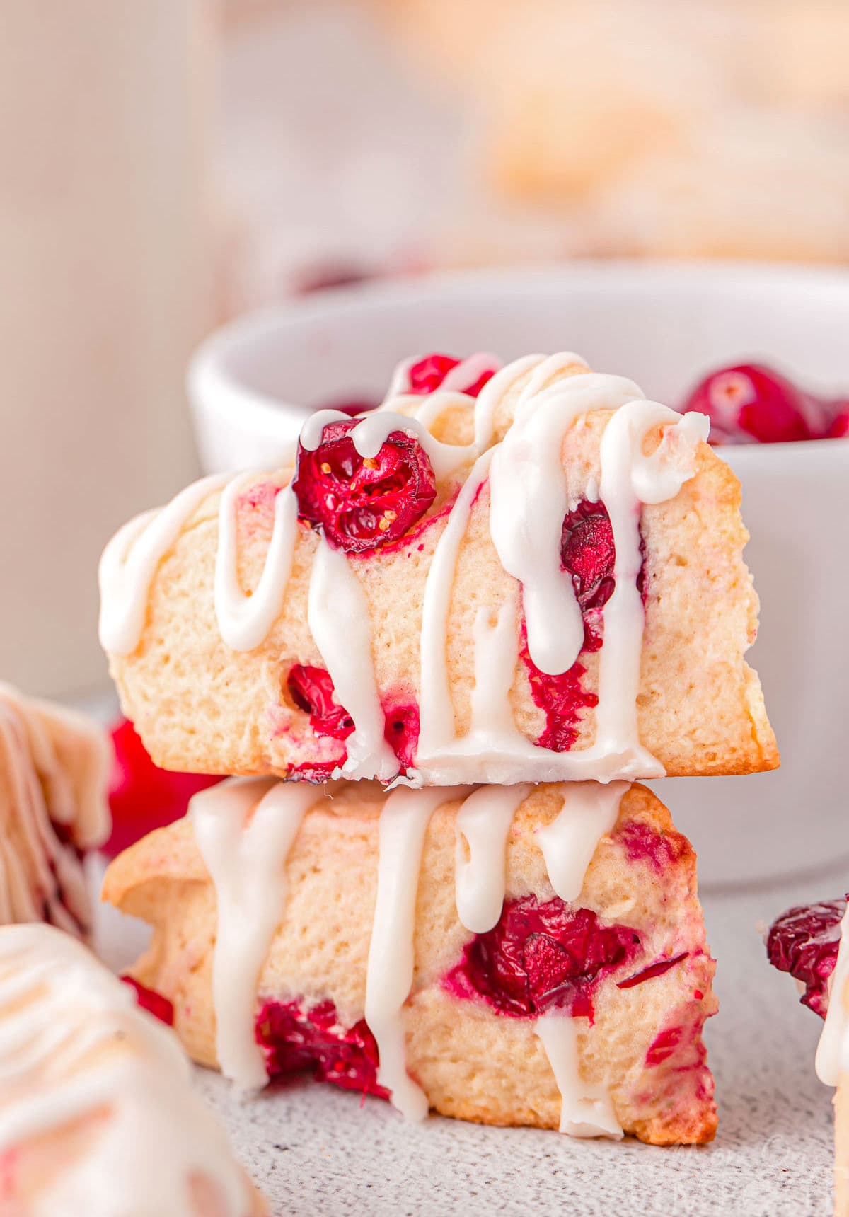 two small cranberry scones stacked with a simple glaze drizzled on top. a small white bowl of cranberries are in back as well as more scones to the sides.