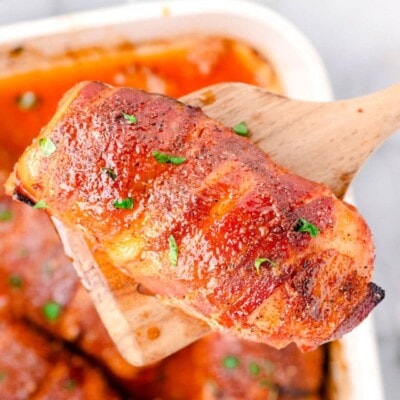 top down look at a casserole dish with bacon wrapped chicken breasts. one piece of chicken is being lifted out of the dish with a wood spoon.