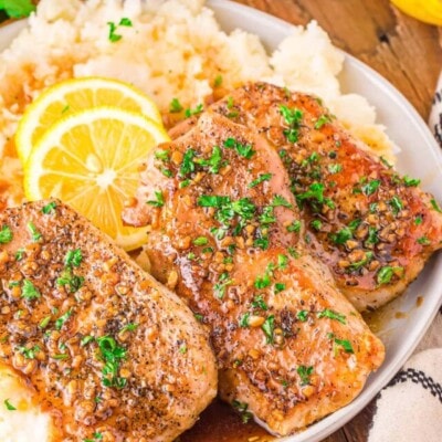 three honey garlic pork chops sitting on a bed of mashed potatoes topped with fresh parsley and lemon slices. plate is on a wood board.