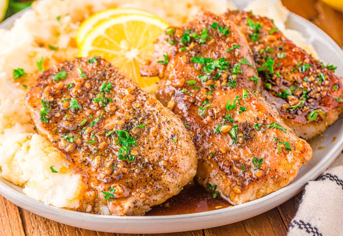 three honey garlic pork chops sitting on a bed of mashed potatoes topped with fresh parsley and lemon slices. plate is on a wood board.