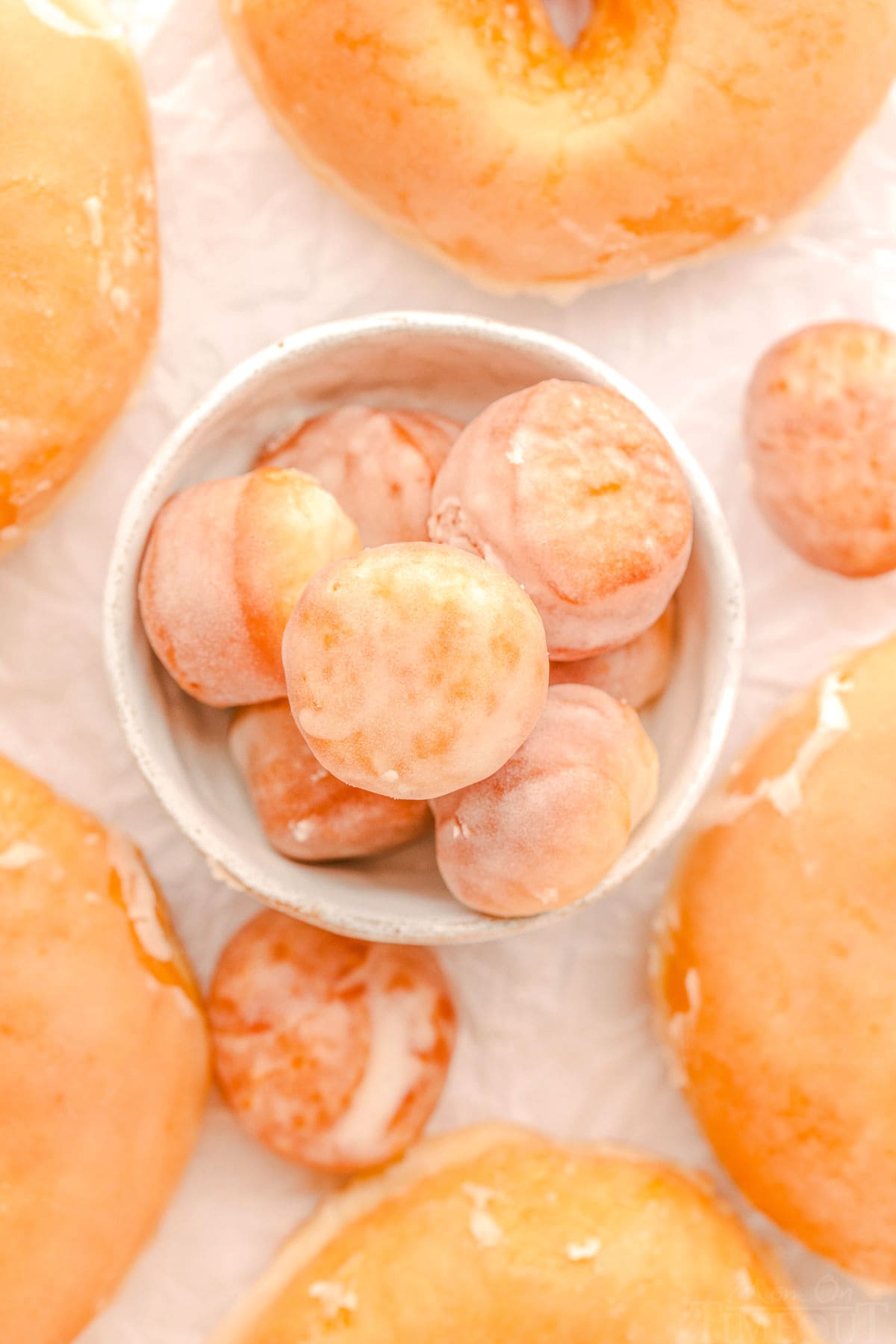 glazed donut holes in a small white bowl top down view. regular donuts surround the bowl.