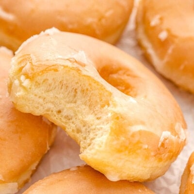 five glazed donuts on white parchment with one donut with a bite taken from it resting at an angle on another donut.