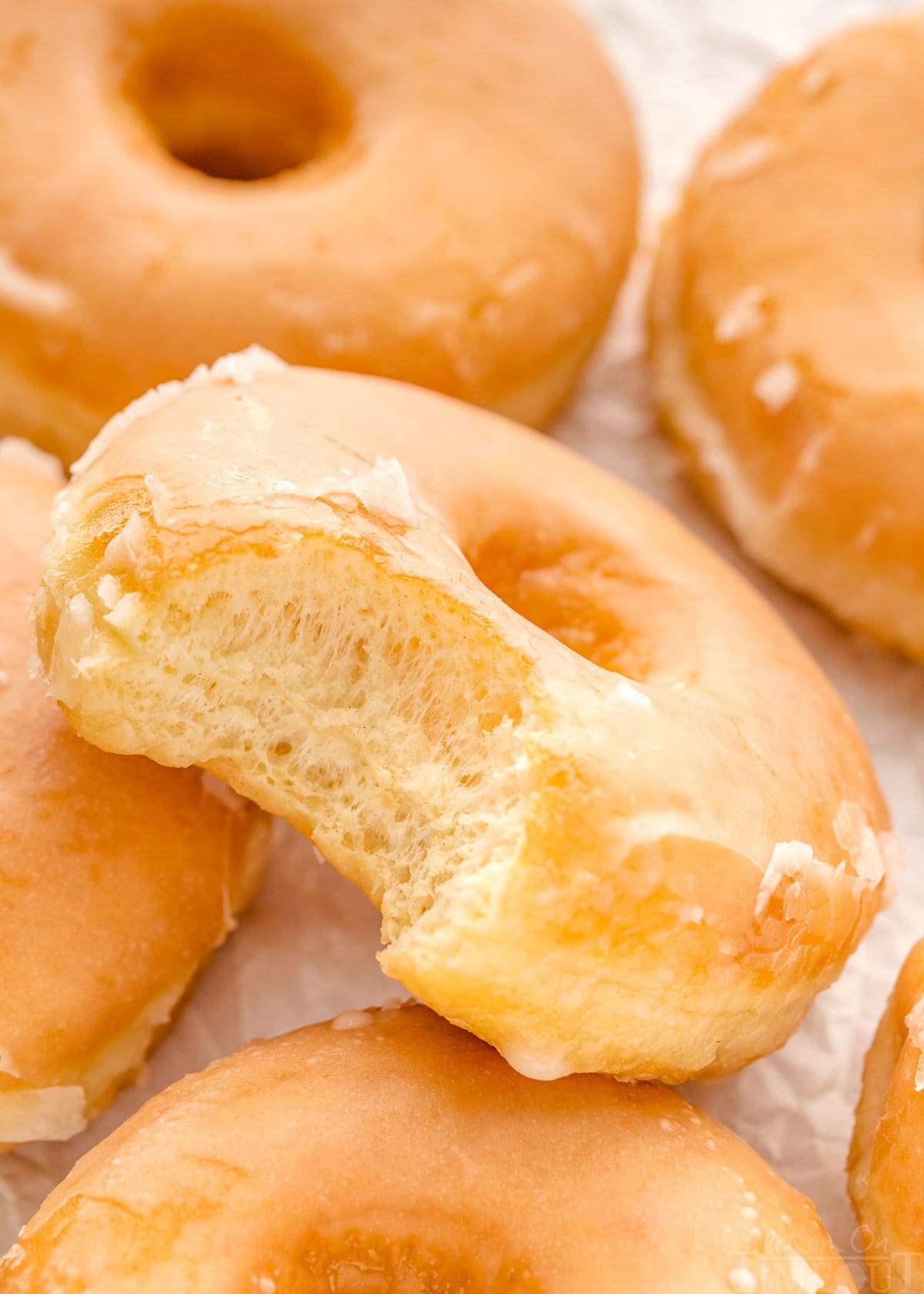 five glazed donuts on white parchment with one donut with a bite taken from it resting at an angle on another donut.