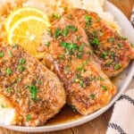 three honey garlic pork chops sitting on a bed of mashed potatoes topped with fresh parsley and lemon slices. plate is on a wood board.