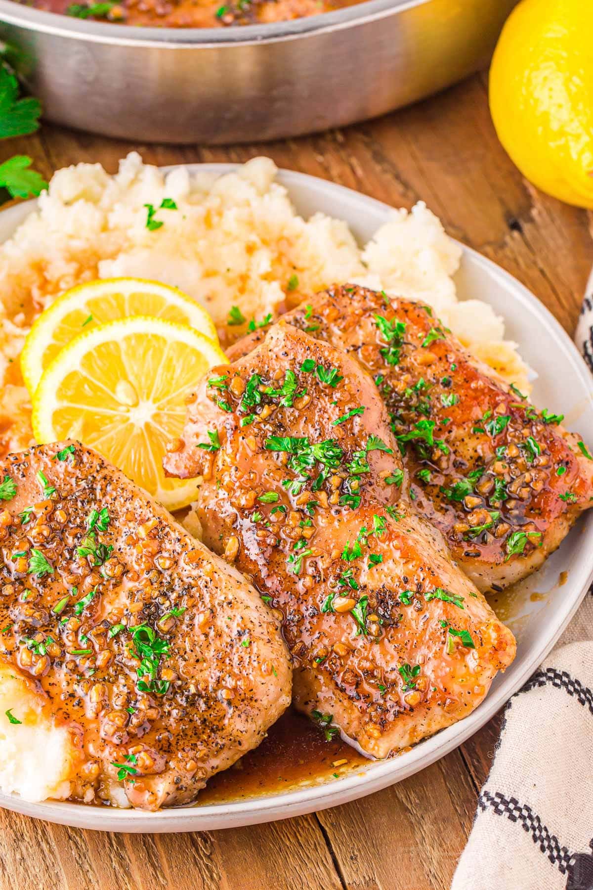 three honey garlic pork chops sitting on a bed of mashed potatoes topped with fresh parsley and lemon slices. plate is on a wood board.