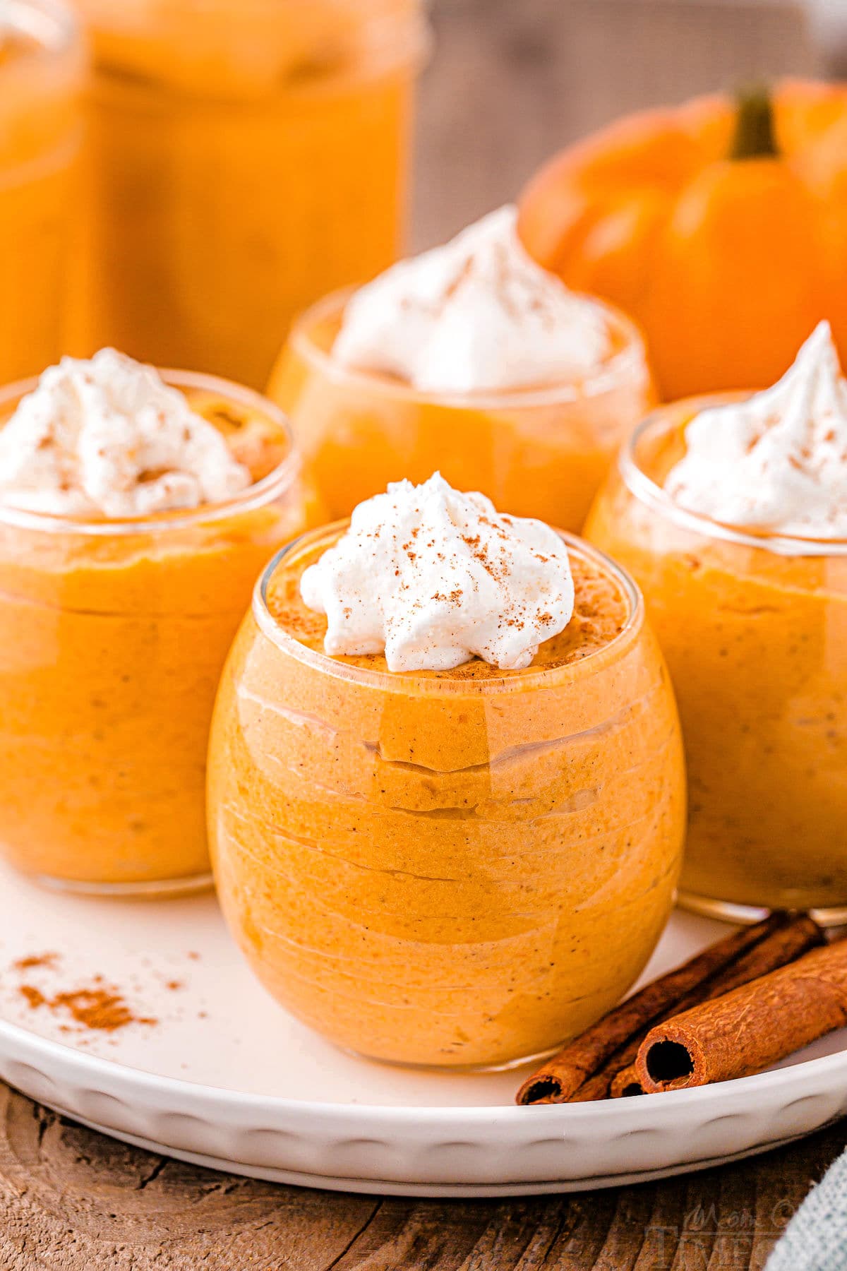 four short wine glasses filled with pumpkin mousse, topped with whipped cream and pumpkin pie spice. the glasses are sitting on a round white plate on a wood surface. there are two cinnamon sticks on the plate.