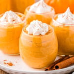 four stemless wine glasses filled with pumpkin mousse, topped with whipped cream and pumpkin pie spice. the glasses are sitting on a round white plate on a wood surface. there are two cinnamon sticks on the plate.