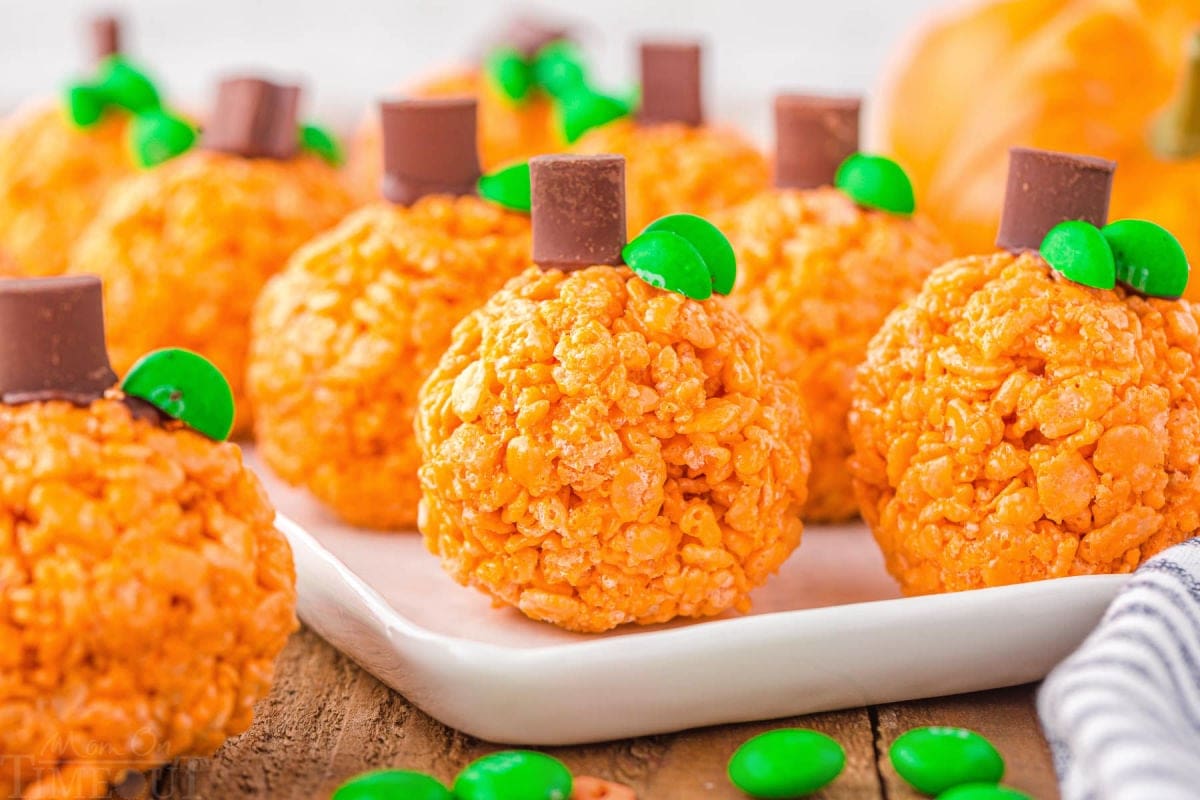 about 10 pumpkin shaped rice krispie treats with chocolate chunk stems and m&m candy leaves sitting on a white platter.