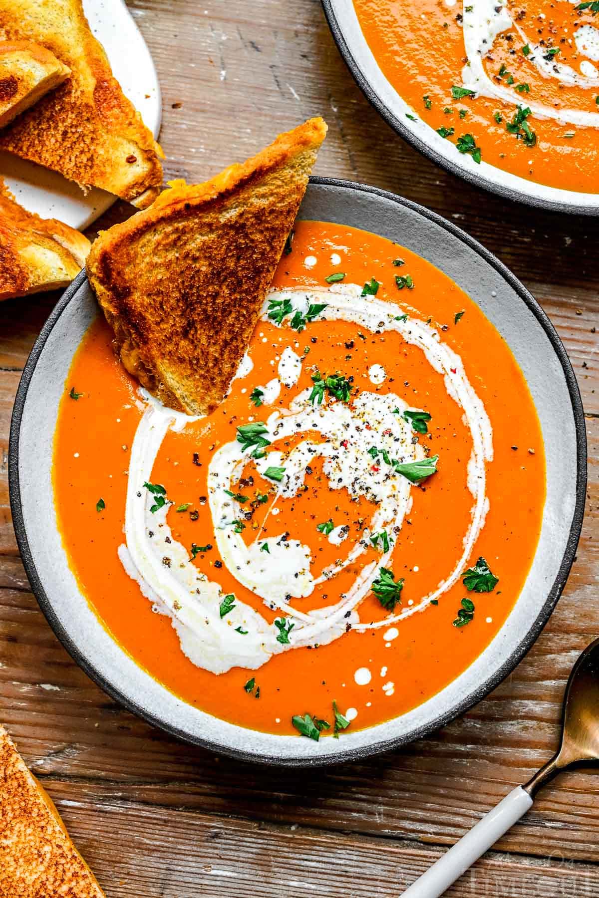 top down view of bowl of tomato soup with half of a grilled cheese sandwich stuck in it. soup has been drizzled with heavy cream, topped with fresh basil and black pepper.